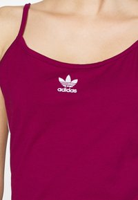 Bordowy top na ramiączkach wykonany z bawełny z cienkimi ramiączkami. Posiada małe białe logo Adidas na klatce piersiowej. Miękka tekstura i casualowy design.