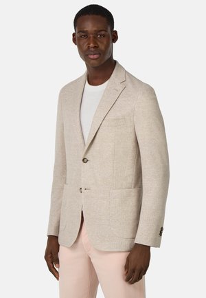 Blazer texturizado beige con dos botones, solapas de muesca y un solo bolsillo en el pecho. Combinado con una camisa blanca y pantalones rosa claro.