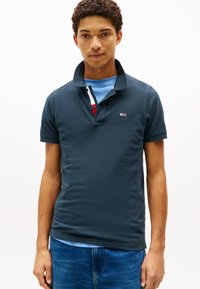 Marineblå poloshirt i teksturert stoff, med klassisk krage, knappestolpe og en logo på venstre bryst. Brukes over en lyseblå t-skjorte.