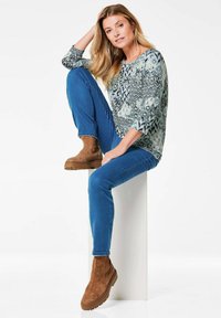 Langärmliges gemustertes Oberteil in Blau, Grün und Weiß; blaue Skinny Jeans; braune Wildleder-Stiefeletten; Modell sitzt auf einem weißen Sockel.