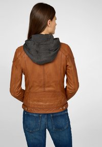 Bruine leren jas met een donkergrijze capuchon, voorzien van gestructureerde naden, subtiele stikdetails en metalen drukknoopsluitingen bij de taille.