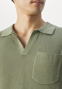 Homme portant un polo en maille texturée vert clair avec une petite poche poitrine et un col ouvert, montré du cou à la poitrine.