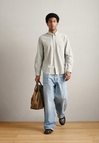 Svetlo siva srajca s gumbi, široke modre jeans hlače, rjavi usnjeni čevlji, nošenje rjave usnjene torbe. Enostavno ozadje, naravna osvetlitev.