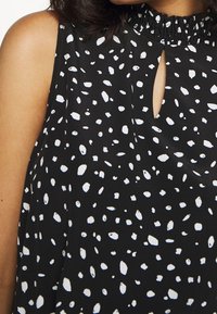 Svart ärmlös topp med vitt prickigt mönster, samlad halsringning och en nyckelhålsdetalj framtill. Slätt, lätt tyg.