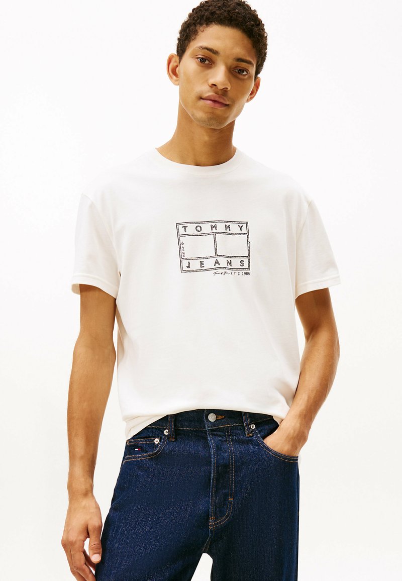 T-shirt bianca in cotone con logo "TOMMY JEANS" in nero, abbinata a jeans di denim blu scuro con un fit classico e dettagli di cucitura sottili.