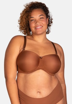 Lächelnde Frau mit lockigem Haar, die einen braunen trägerlosen BH und passende Unterwäsche vor hellem Hintergrund präsentiert.