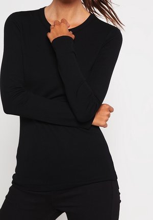 Femme portant un chemisier noir moulant à manches longues, les bras croisés avec une main touchant son cou, sur un fond clair uni.