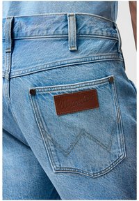 Jasnoniebieskie dżinsy charakteryzują się teksturowaną tkaniną dżinsową, tylną kieszenią z brązowym skórzanym naszywkiem z wytłoczonym napisem "Wrangler" oraz detalami w kontrastowym przeszyciu.