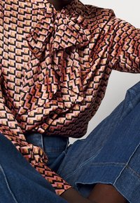 Silke skjorte med geometrisk mønster i pink, sort og guld. Har en stor sløjfekrave og flæset foran, parret med denim jeans.