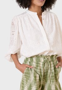 Blusa bianca ricamata con colletto alto, maniche a sbuffo e chiusura a bottoni, abbinata a pantaloni verdi con motivo e coulisse.