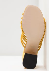 Keltainen mokkanahkainen korkokenkä, jossa on useita pehmeitä hihnoja ja teksturoitu musta kumipohja. Sileä vaaleanruskea sisäpohja; valmistajan tiedot näkyvissä.