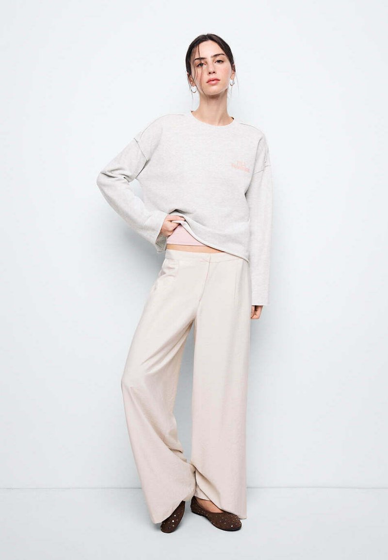 Jeune femme portant un pull ample gris clair et un pantalon large beige, debout avec une main dans la poche sur un fond blanc uni.