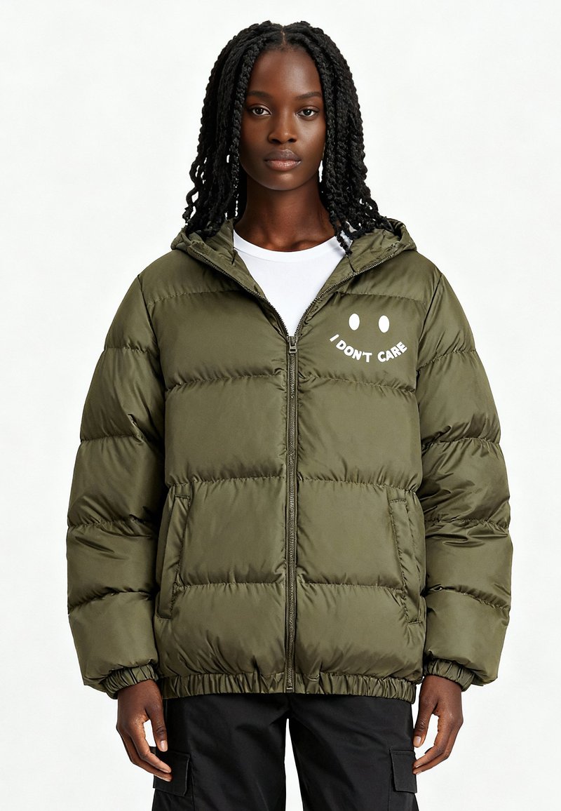 Doudoune vert olive avec fermeture éclair à l'avant, poignets élastiques et un motif brodé "JE M'EN FOUS". Comprend une capuche et des poches latérales.
