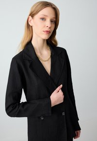 Czarny żakiet z lnu z podwójnym zapięciem, klapami o prostokątnym kształcie i guzikami. Noszony na warstwowym naszyjniku z złotych łańcuszków, o gładkiej strukturze.