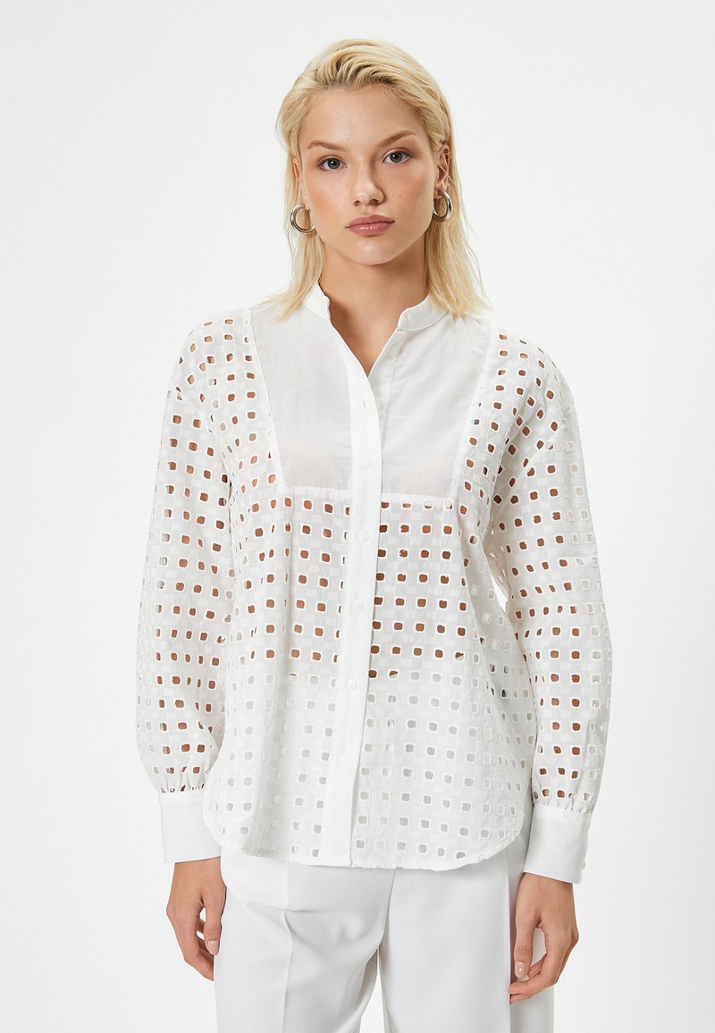 Lyshåret kvinde iført en hvid skjorte med knapper og et firkantet udskæringmønster samt hvide bukser, står mod en ensfarvet baggrund.