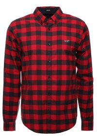 Camisa de franela a cuadros rojos y negros con cuello abotonado, material elástico y un pequeño logo en el lado izquierdo del pecho.