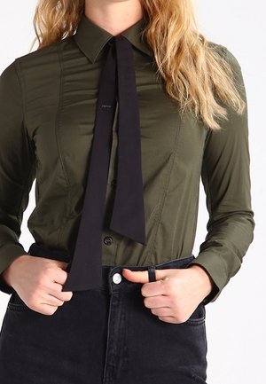 Kobieta w oliwkowozielonej koszuli zapinanej na guziki z czarnym krawatem, wkładająca ręce do kieszeni czarnych, wysokiej talii jeansów.