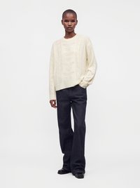 Personne mince aux cheveux courts portant un pull en maille crème avec un motif de feuilles, un pantalon noir ample et des chaussures noires, debout devant un fond uni.