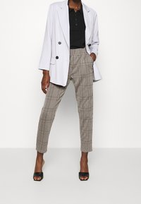 Bílý dvouřadý blejzr přes černé polo, doplněný hnědými kostkovanými kalhotami a černými sandály na podpatku s otevřenou špičkou. Plochá textura.