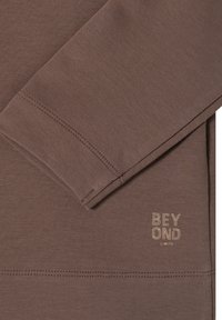 Brązowa bluzka z długim rękawem wykonana z teksturowanej tkaniny o dopasowanym fasonie. Logo "BEYOND LIMITS" w jasnym złotym kolorze umieszczone blisko dołu.
