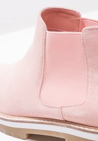 Różowe zamszowe botki za kostkę z elastycznymi panelem bocznymi, teksturowanym wykończeniem oraz białą podeszwą środkową i jasnobrązową podeszwą zewnętrzną.