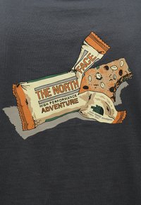 Γραφική απεικόνιση περιτυλίγματος μπάρας σνακ και μιας μερικώς δαγκ ωμένης μπάρας, με χρώματα μπεζ, πορτοκαλί και πράσινο,που φέρει το λογότυπο "The North Face".