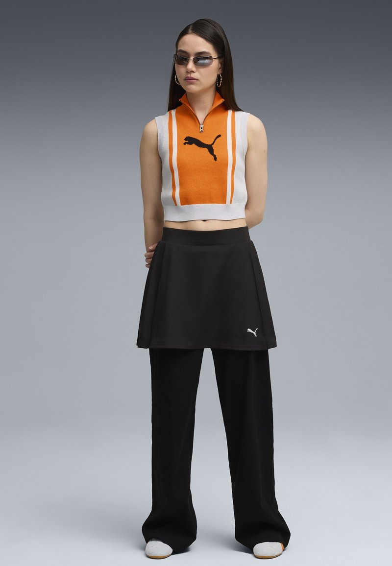 Suéter corto naranja y blanco con cremallera, con el logo de Puma en negro; combinado con una falda negra de corte acampanado y pantalones negros de pierna ancha.