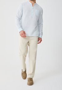 Uomo che cammina indossando una camicia a maniche lunghe blu chiaro, pantaloni beige e mocassini di camoscio marrone su sfondo bianco semplice.