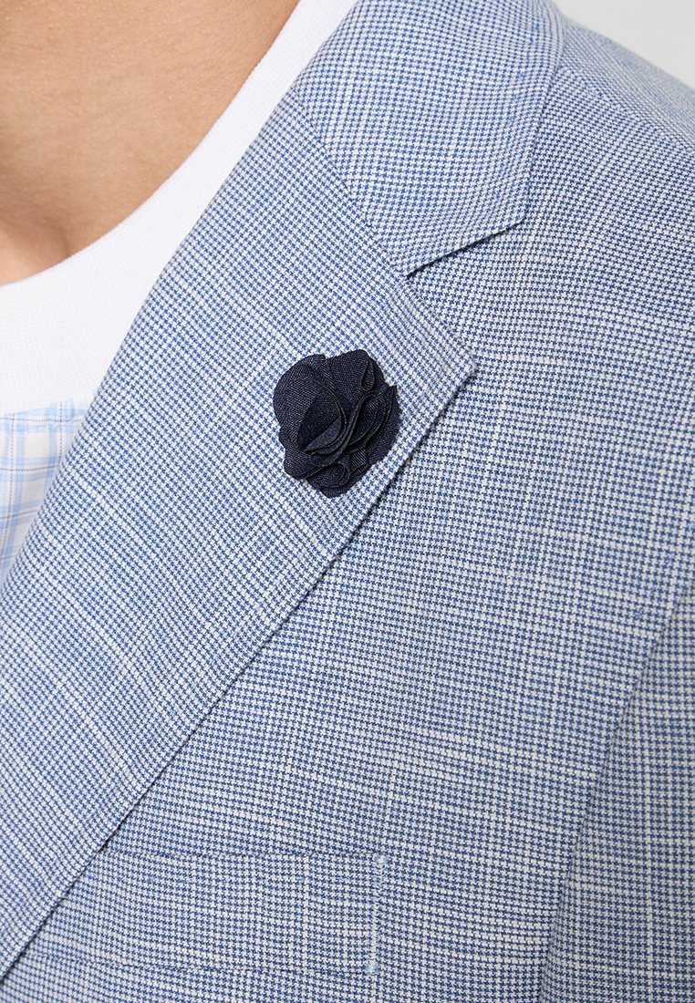 Blazer a quadri blu chiaro con una spilla a forma di fiore in tessuto blu navy sul bavero. Tessuto strutturato con un motivo sottile, tasca singola sul davanti della giacca.
