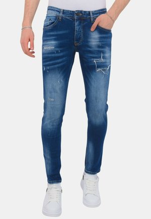 Man draagt een slim-fit blauwe versleten spijkerbroek met vervaagde details en witte sneakers, staand met één hand in de zak.