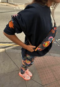 Musta collegepaita, jossa on värikäs auringon kirjailu, kukkakuviolliset leggingsit ja persikanväriset lenkkarit. Asusteena musta vyölaukku, jossa on eloisat yksityiskohdat.