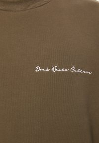 Brun bomull genser med høy hals og hvit brodert tekst "Don't Waste Culture" på forsiden. Jevn tekstur, uformell passform.