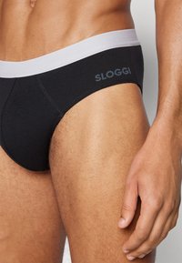 Schwarze Baumwollslips mit einem hellgrauen elastischen Bund. Mit SLOGGI-Branding am Bund und einem glatten, figurbetonten Design.