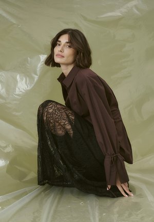 Camicia marrone oversize con maniche ampie, abbinata a una gonna in pizzo nero trasparente. Sullo sfondo c'è una plastica verde chiaro sgualcita.