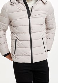 Světle šedá prošívaná bunda s předním zipem, černými detaily a kapucí. Má vodorovné šití a boční kapsy.
