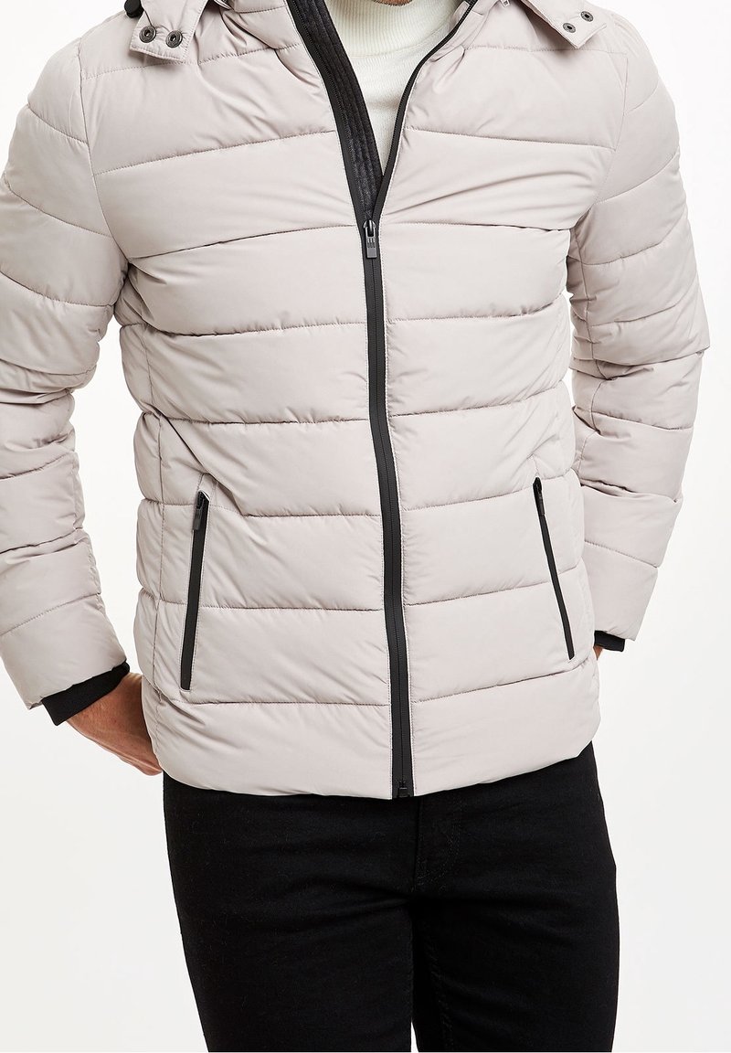 Světle šedá prošívaná bunda s předním zipem, černými detaily a kapucí. Má vodorovné šití a boční kapsy.