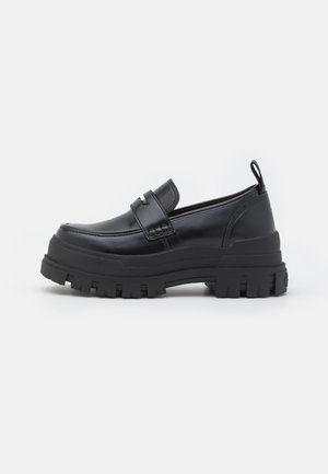 Musta nahkainen loafer-kenkä, jossa paksu uurrettu pohja, penny-hihna ja vetokiristin kantapäässä, sivulta kuvattuna valkoisella taustalla.
