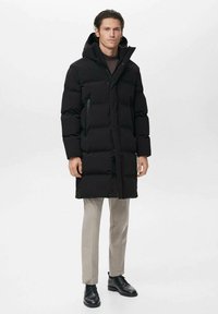 Abrigo acolchado negro con capucha, que cuenta con un bolsillo con cremallera en el lado izquierdo del pecho. El modelo lleva pantalones beige y zapatos negros, de pie frente a un fondo liso.