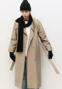 Manteau beige à double boutonnage, avec des liens de ceinture, superposé à une veste blanche zippée, complété par une écharpe noire et un bonnet.