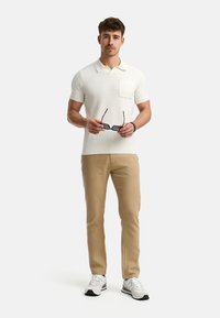 Camisa polo blanca de canalé con mangas cortas y bolsillo, combinada con pantalones color canela y zapatillas blancas con acentos grises.