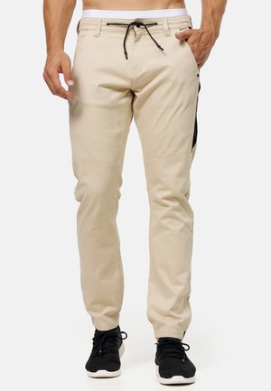 Pantalones ligeros beige con cintura con cordón, ajuste ajustado, bolsillos laterales y acentos negros a los lados. Los lleva puestos con zapatillas negras.