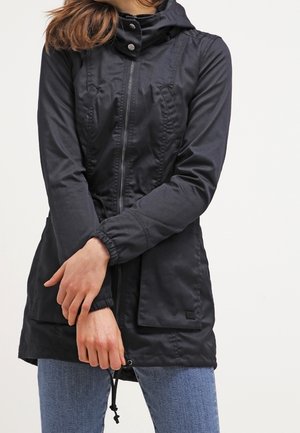 Kobieta w długiej czarnej kurtce na zamek z wysokim kołnierzem i niebieskich dżinsach, ręce skrzyżowane na wysokości talii.