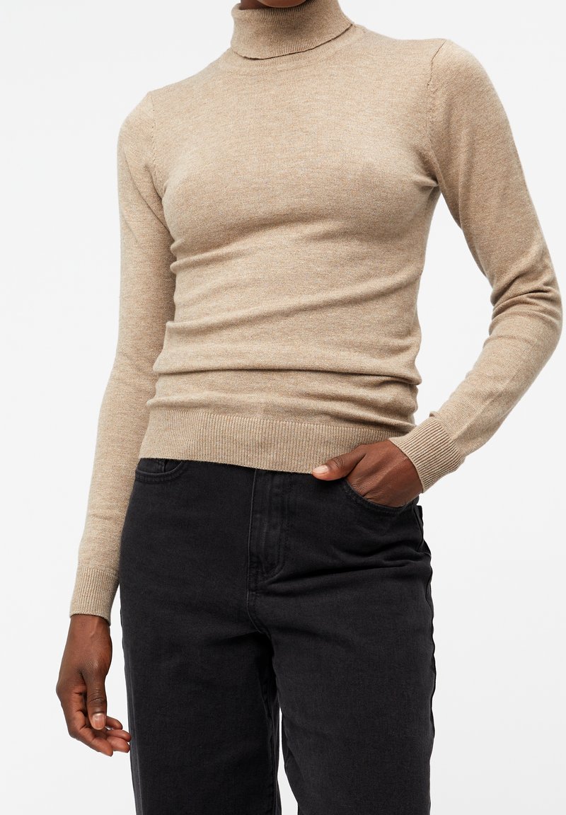 Pull en col roulé beige, design ajusté, poignets et ourlet côtelés, texture douce. Assorti avec un pantalon en denim noir.