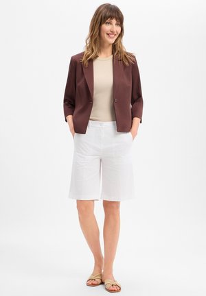 Glimlachende vrouw staand met handen in de zakken, draagt een bruine blazer, beige top, witte knielange shorts en beige sandalen op een witte achtergrond.