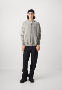 Siva zip-up kapuca s logotipom, sprednjimi žepi in rebrastimi manšetami, v kombinaciji s črnimi cargo hlačami in črnimi supergami. Model nosi črno kapico.