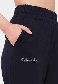 Navy blue cotton jogger pants with an elastic waistband, side pockets, and embroidered “C Sports Club” logo in white. Smooth texture.