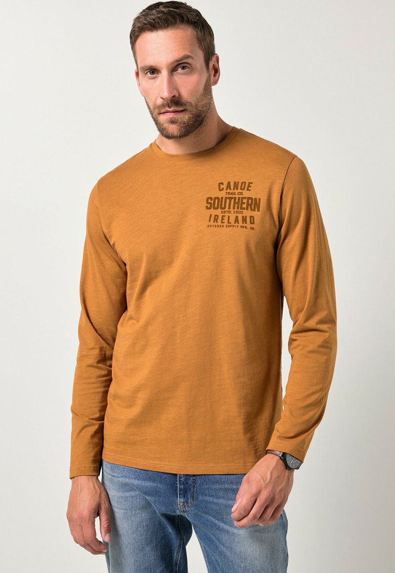Langarmige mosterdkleurige blouse met een gestructureerde afwerking, voorzien van een donkere gedrukte logo op de linkerborst en gecombineerd met denim jeans.