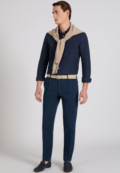 Man wearing navy shirt and trousers with beige sweater draped over shoulders, beige belt, and dark loafers, standing against plain background.