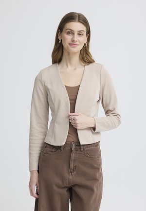 Jeune femme aux cheveux châtain clair portant une veste beige ouverte devant, un top marron et un pantalon marron taille haute, debout devant un fond uni.