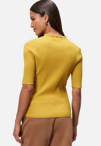 Gele geribde trui met korte mouwen, heeft een golvende halslijn en textuurstof. Gekozen in combinatie met bruine broek, achterkant zichtbaar.